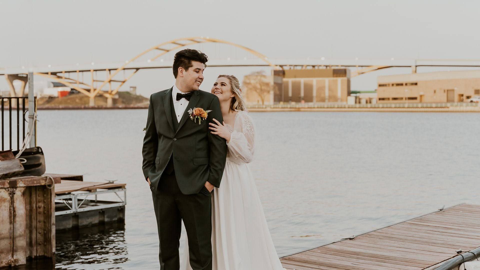 Couple in formal attire standing by a waterfront dock with a bridge in the background