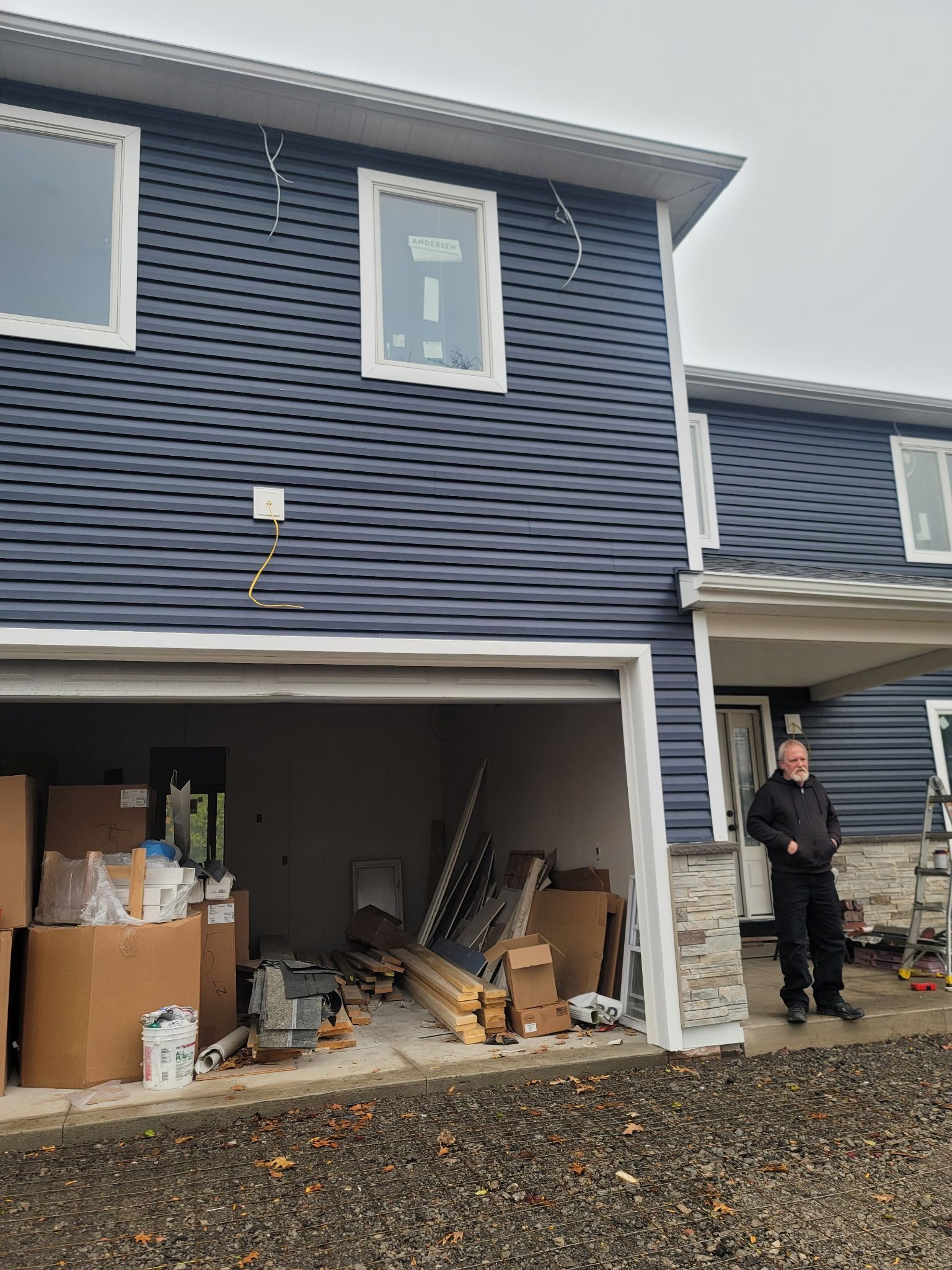 A person stands outside a new house with blue siding and an open garage containing construction materials and tools.