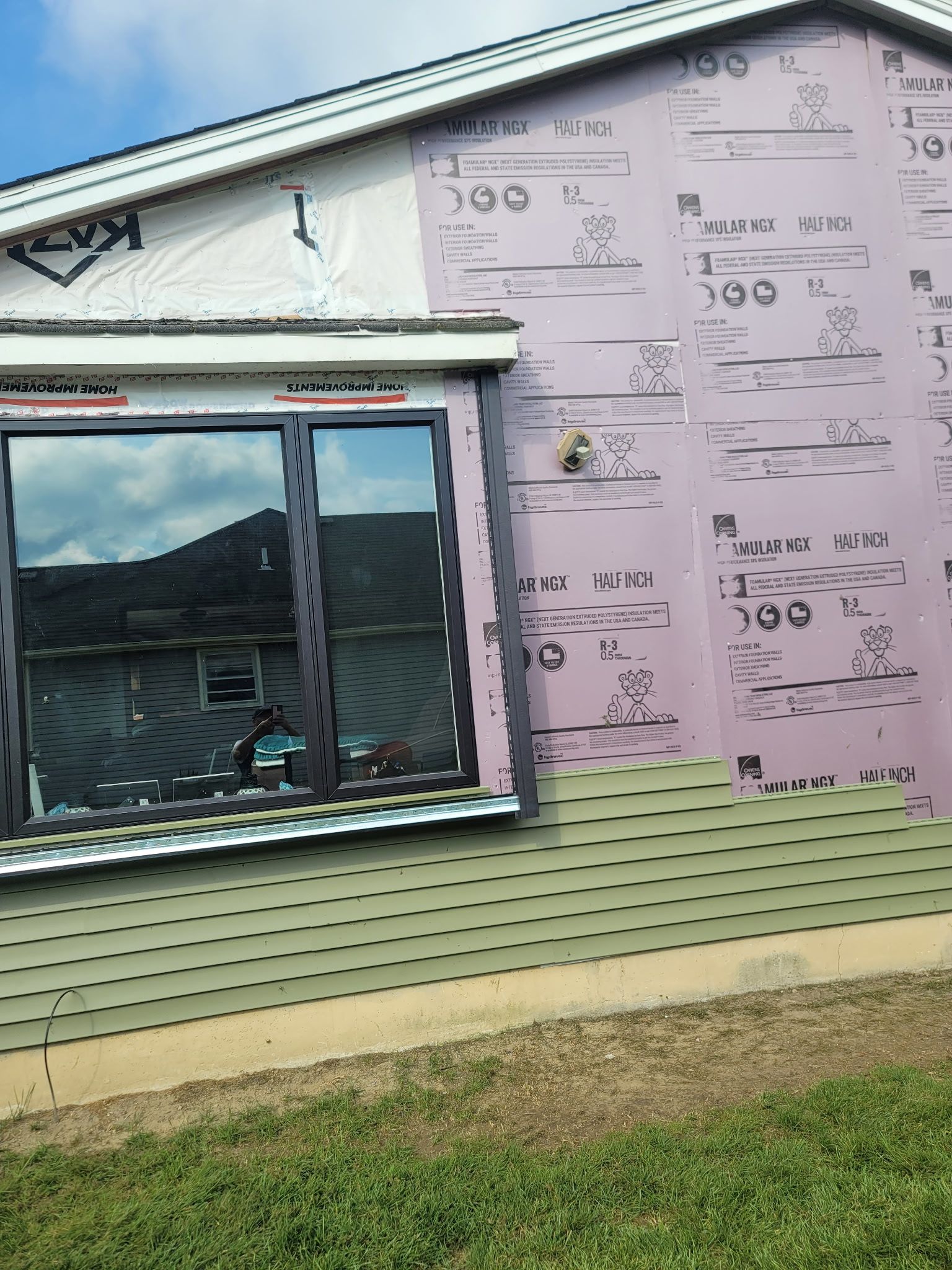 Exterior wall of a house under renovation with light green vinyl siding, purple foam insulation, and a dark window frame.