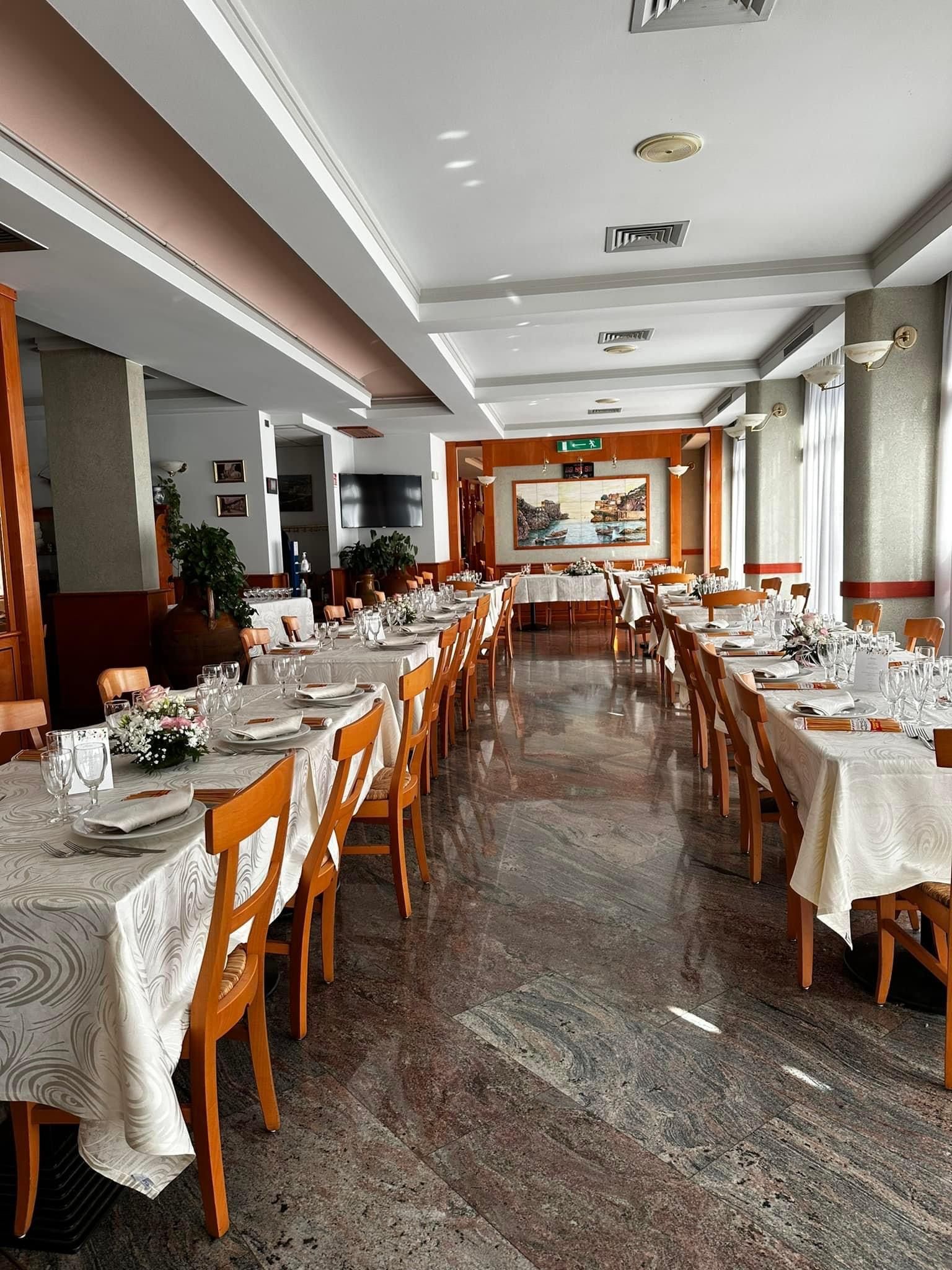 Lungo interno di un ristorante con file di tavoli apparecchiati con tovaglie bianche, pronti per i commensali.