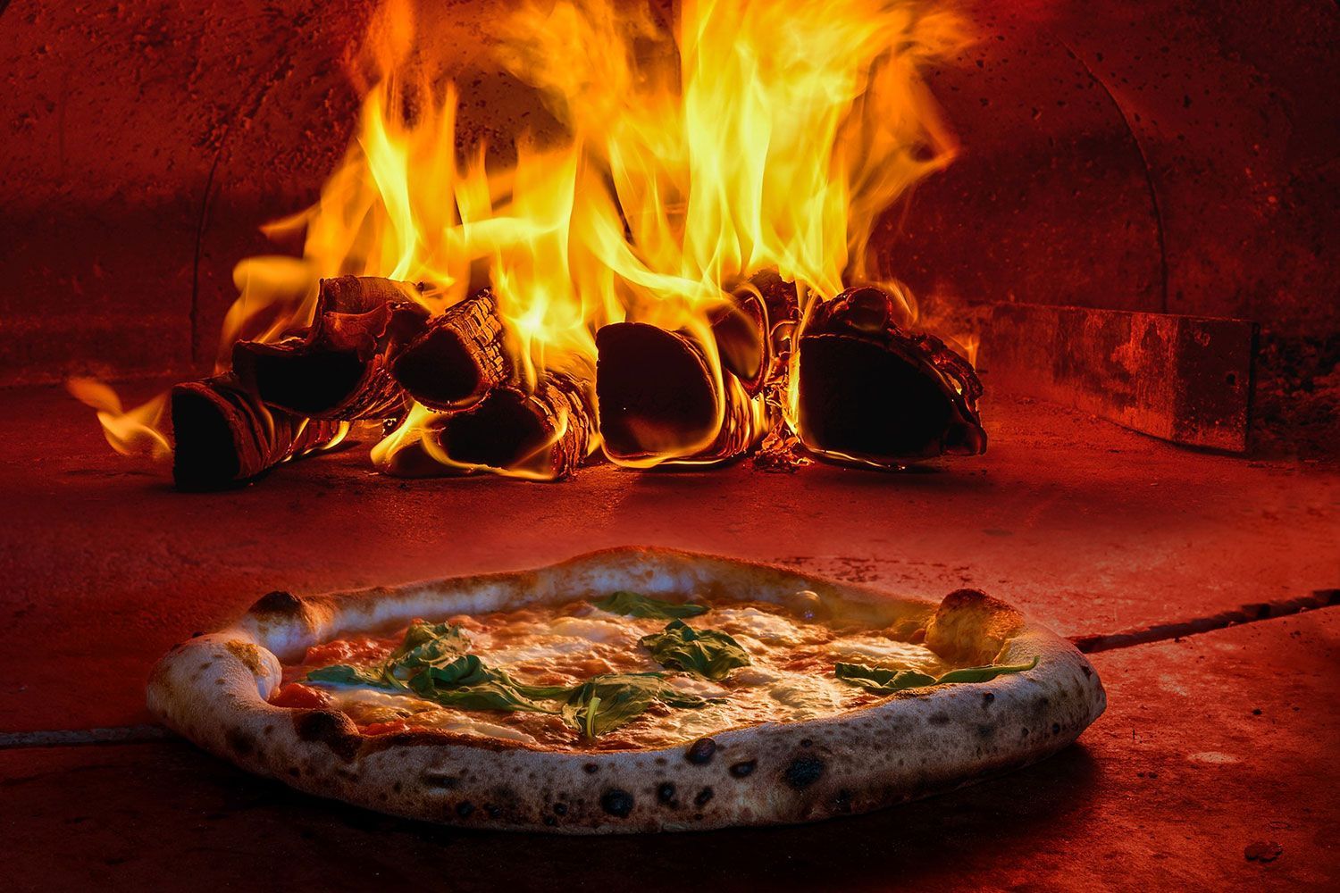 Pizza cotta nel forno a legna con fiamme sullo sfondo.