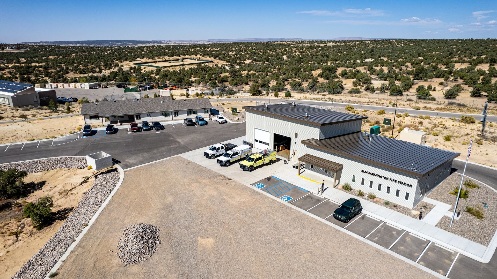 Wide aerial view of a fire station with dark metal roofs, parked vehicles and a gravel lot.