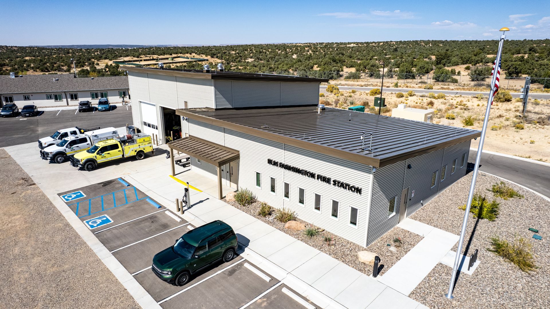 Angled aerial view of a gray fire station with dark metal roofs, garage bays and marked parking spaces.