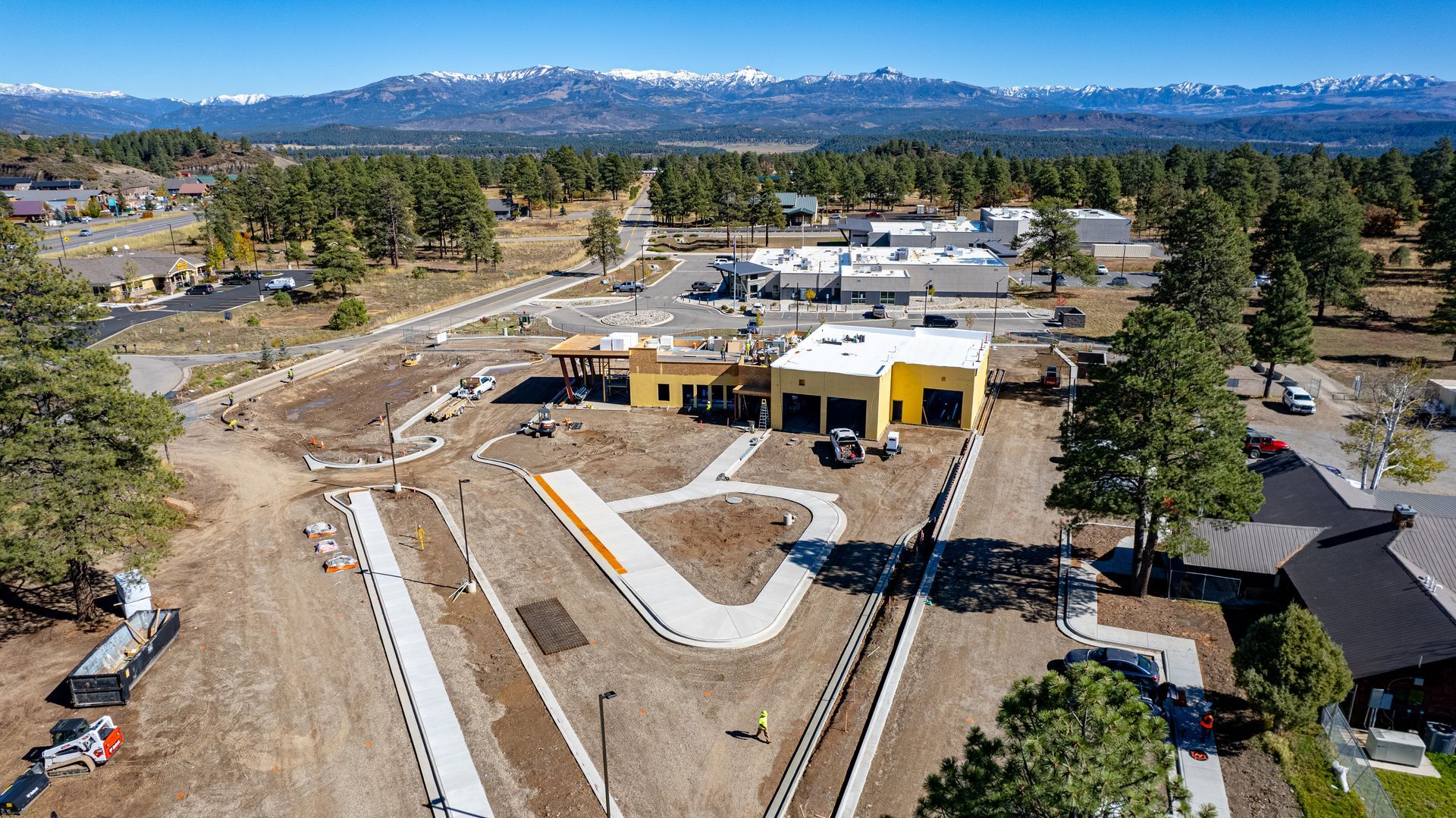 Wide aerial view of a construction site with a white-roofed building, new curbs and surrounding pine trees.