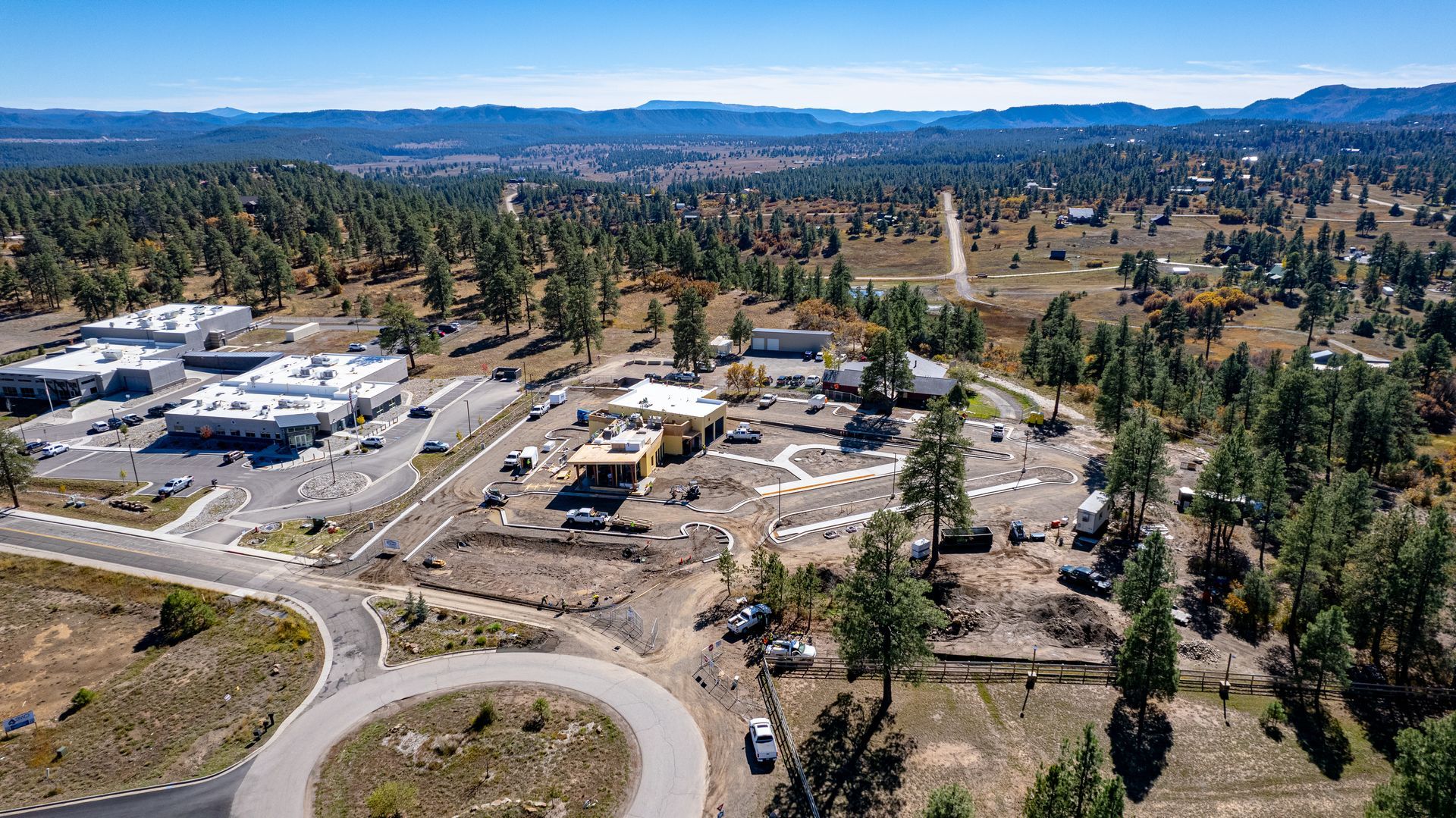 Wide aerial view of a building construction site near a roundabout, roads and wooded hills.