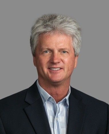 Professional headshot of a smiling person in a dark blazer and light blue shirt on a gray background