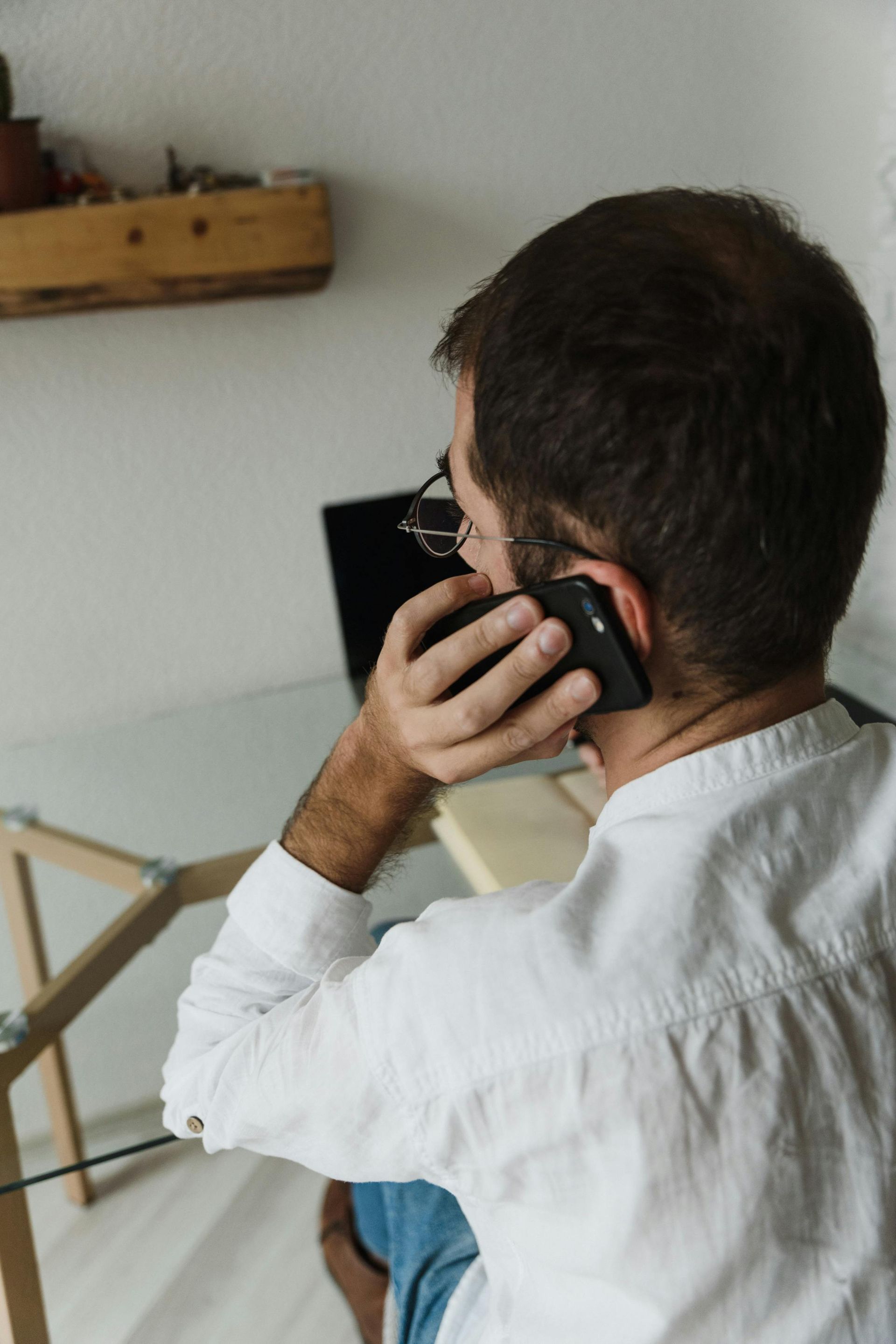 Man seated indoors, holding a phone to his ear while facing a laptop on a table.