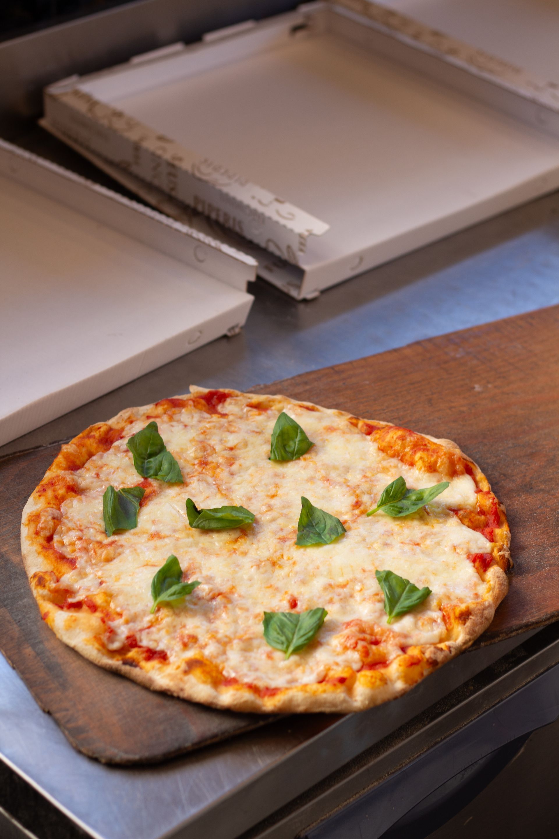 Pizza su un tagliere di legno, condita con formaggio e basilico, vicino a vassoi di metallo.