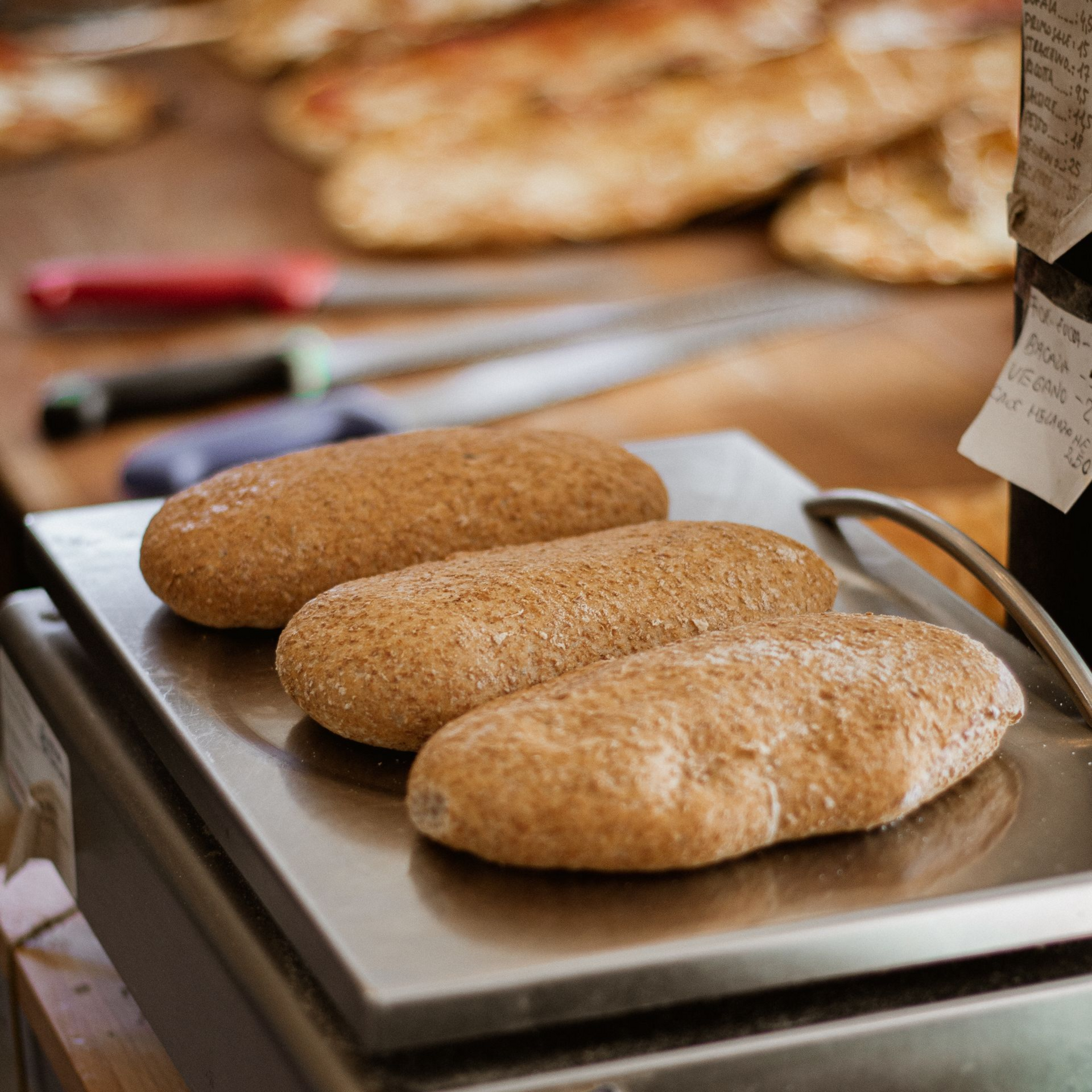 Tre pagnotte di pane su una bilancia in un panificio, con altre pagnotte e coltelli sullo sfondo.