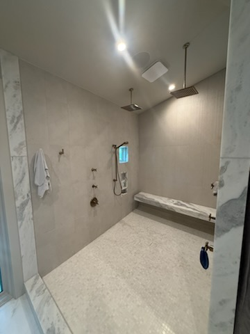 Spacious shower with two showerheads, bench, and various fixtures; light gray walls and white marble accents.