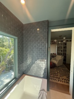 Bathroom with blue-tiled wall, window, and a door leading to a room with a rug and armchair.