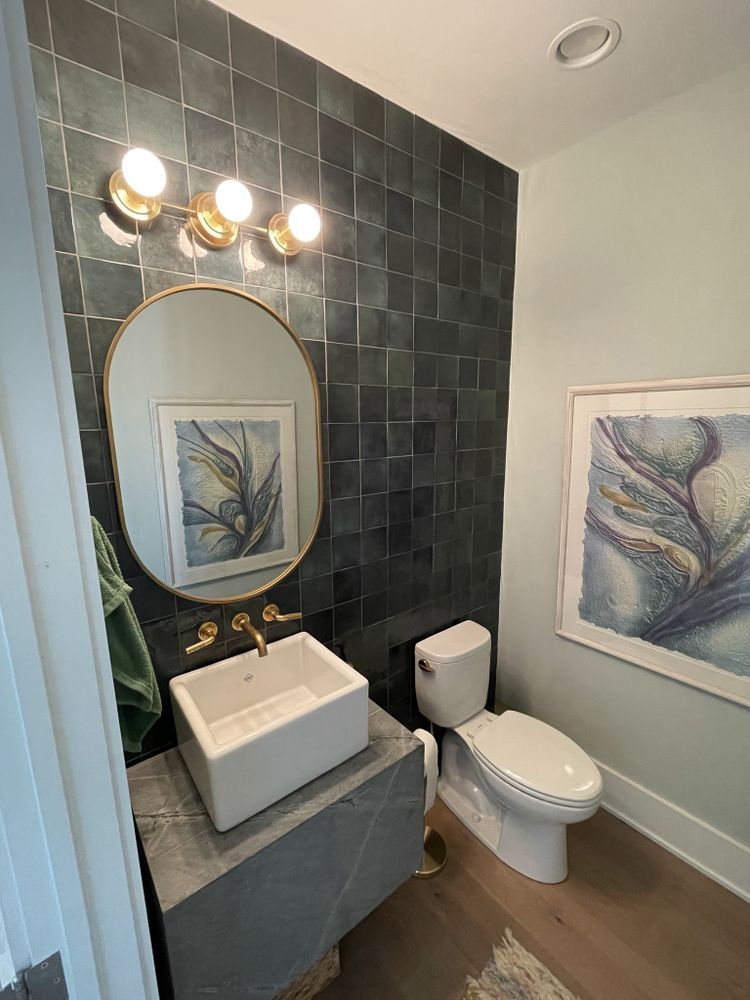 Bathroom with dark tiled accent wall, gold fixtures, oval mirror, and artwork.