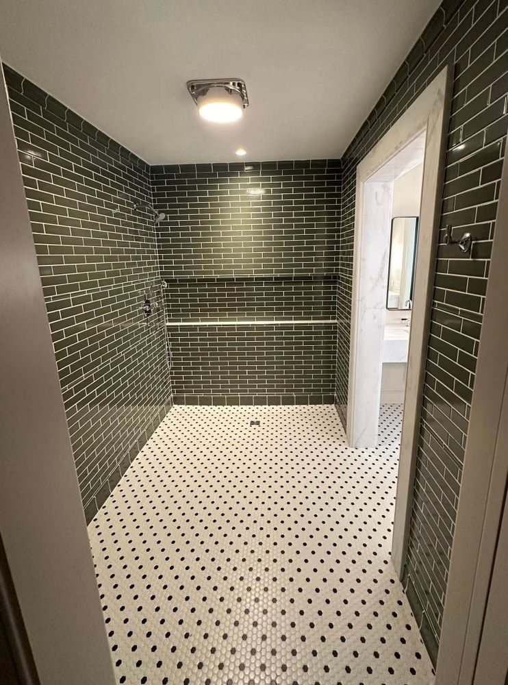 Walk-in shower with dark green brick-like tile walls, white and black mosaic tile floor, and open doorway to a bathroom.