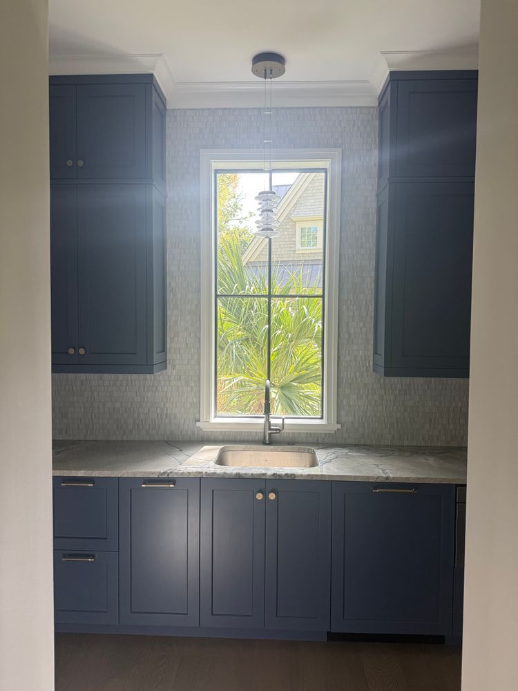 Navy cabinets flank a window above a sink in a kitchen, neutral-colored backsplash, light fixture.