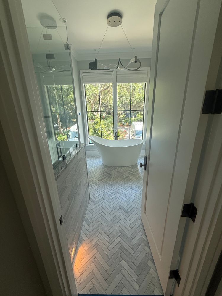 Bathroom with herringbone tile floor, white tub in front of window, marble wall, seen from doorway.