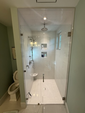 Shower stall with glass doors, white tiled walls and floor, stainless steel fixtures, and a toilet.
