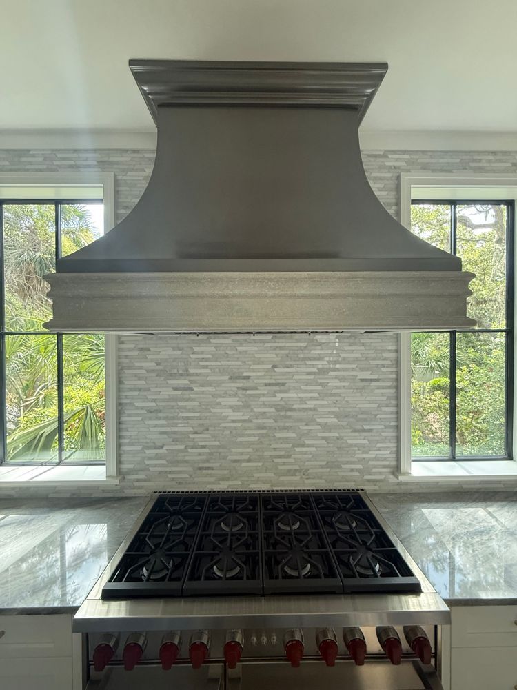 Kitchen with stainless steel range and hood, gray stone tile backsplash, and two windows with a green outdoor view.