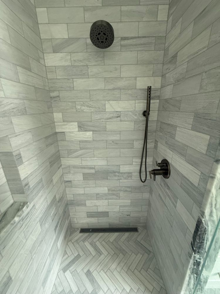 Shower with light gray brick-patterned tiles, dark showerhead, and a drain.