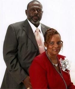 A man in a gray suit and a woman in a red blazer with a floral corsage pose together against a white background.