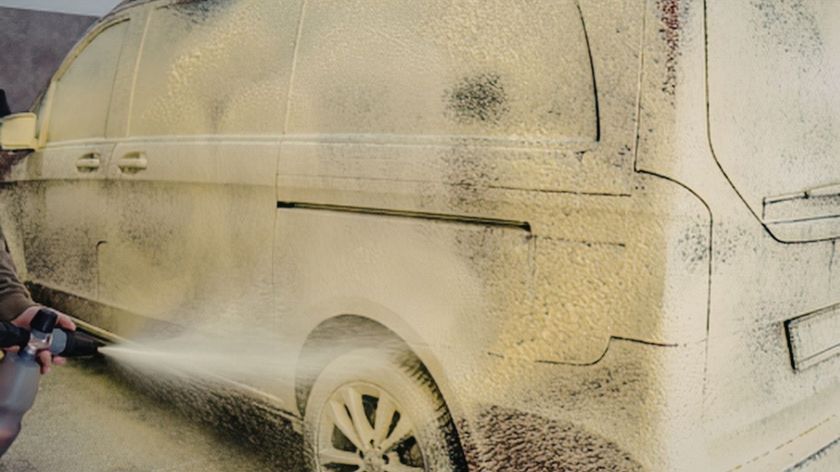 A pressure washer sprays thick, yellow cleaning foam onto the side of a parked van.