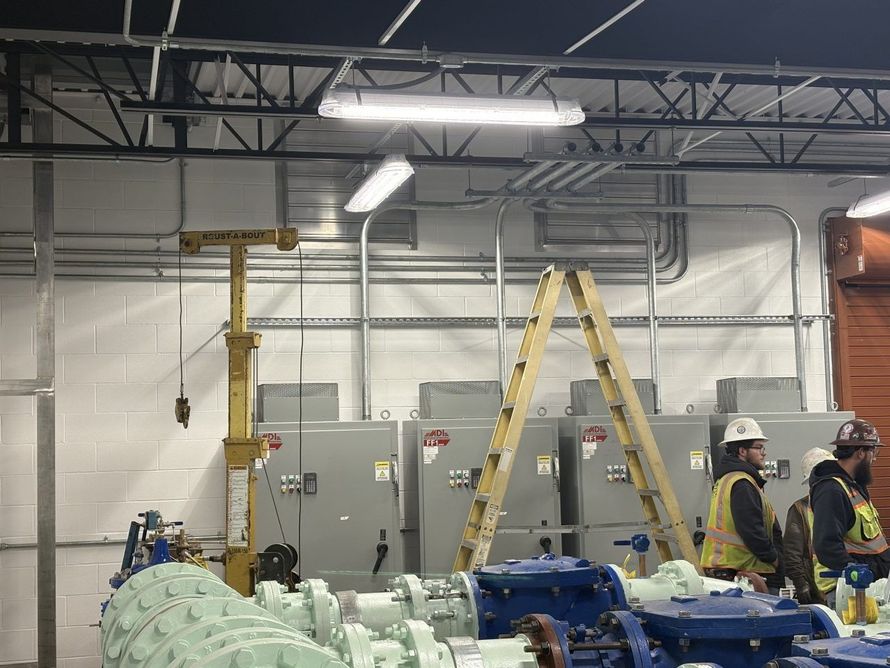 Construction workers in hard hats and vests work near large electrical panels and blue industrial machinery in a warehouse.
