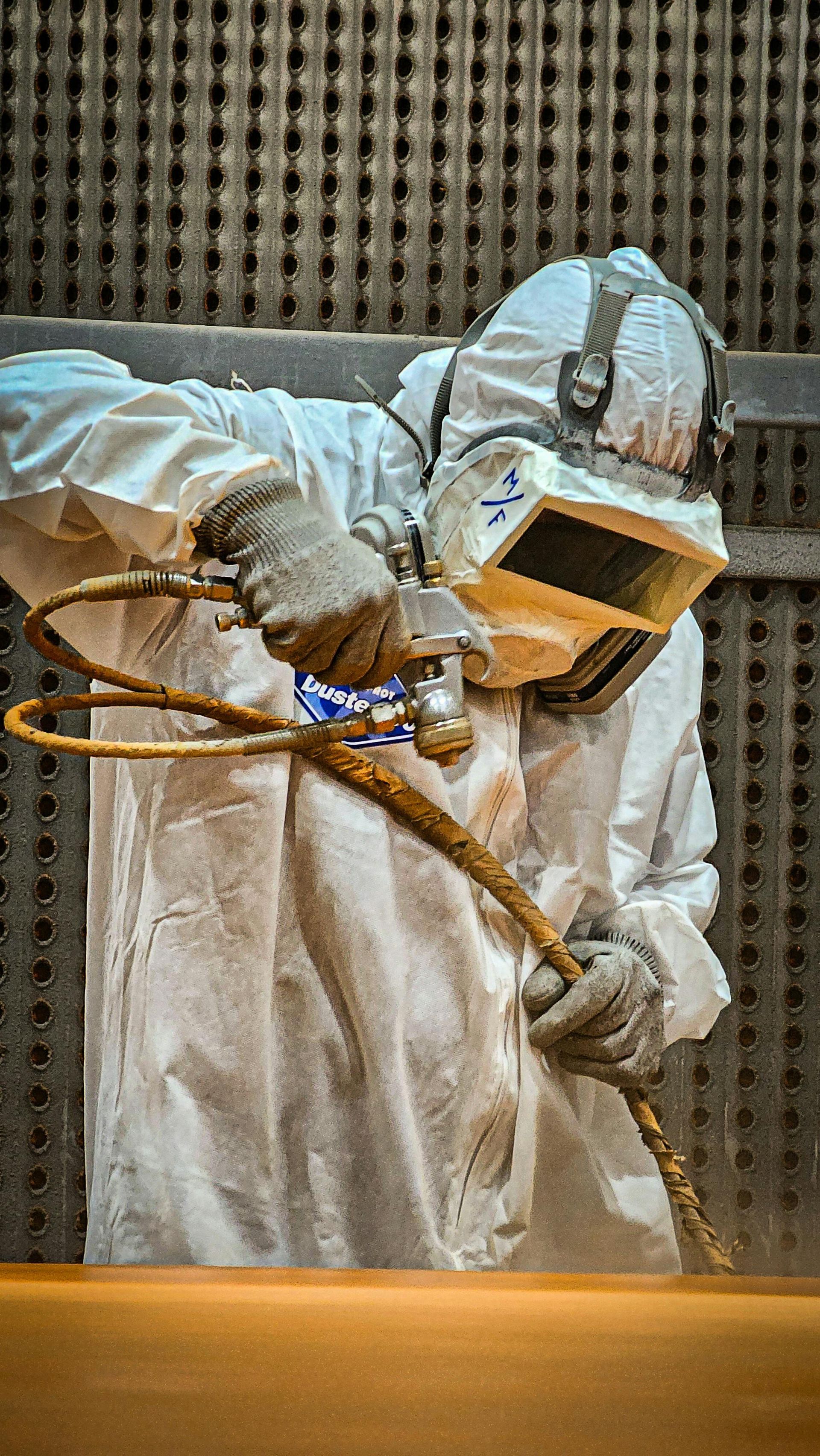 A worker in a white protective suit and helmet uses a spray gun in an industrial facility.