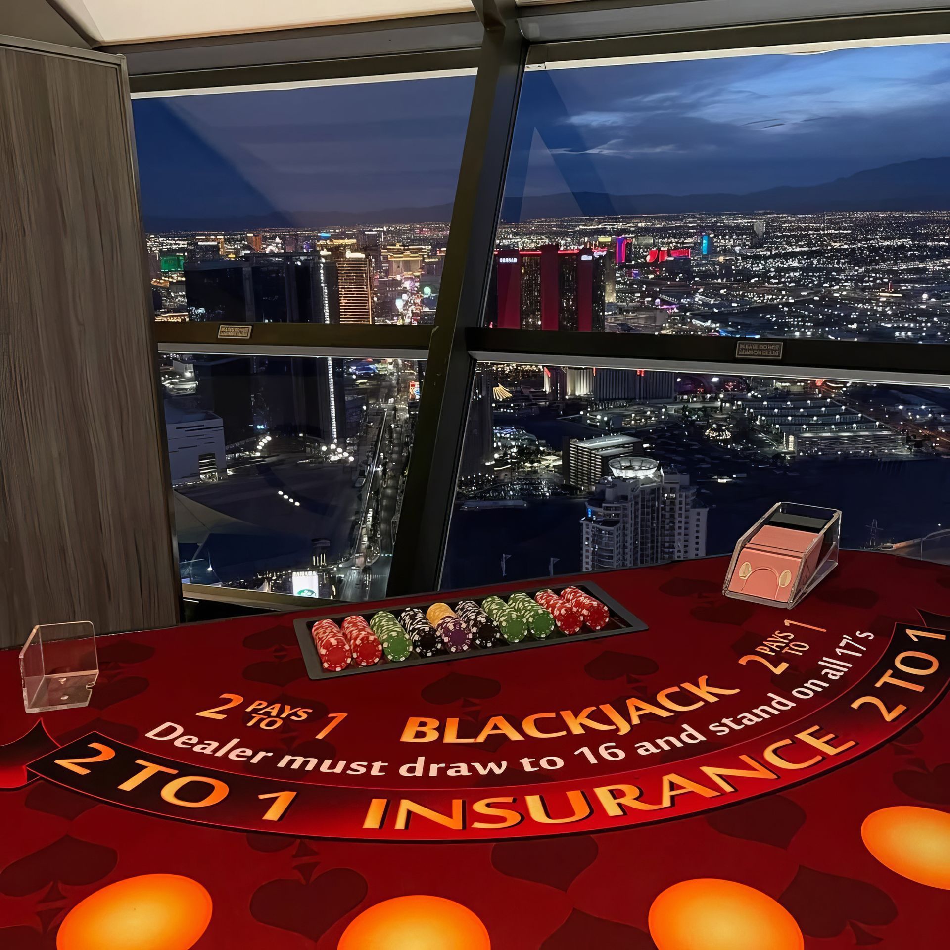 Blackjack table by a window overlooking a city skyline at night, with cards and chips on red felt.