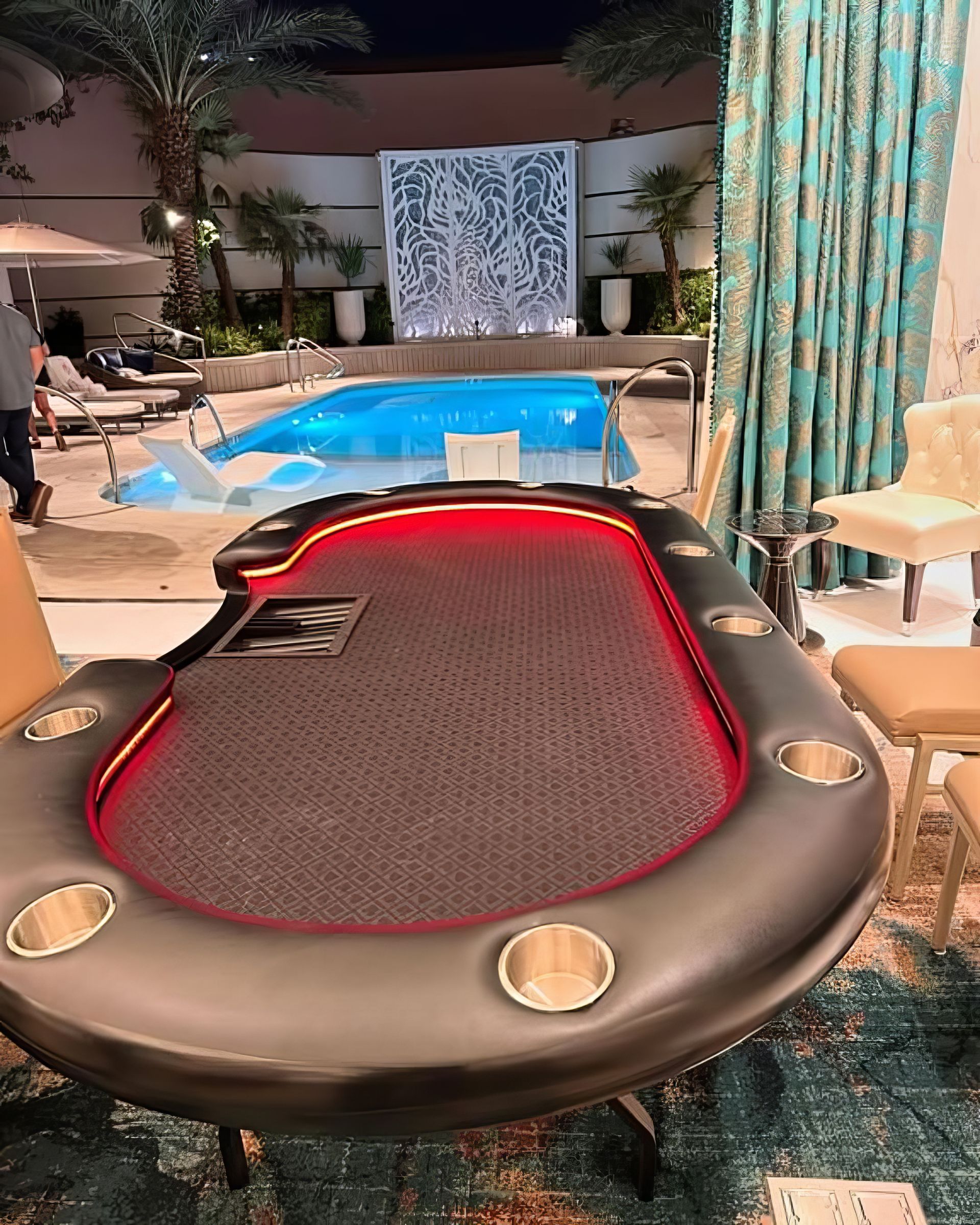 Outdoor gaming table with red trim beside a pool at night, surrounded by lounge chairs and palm trees