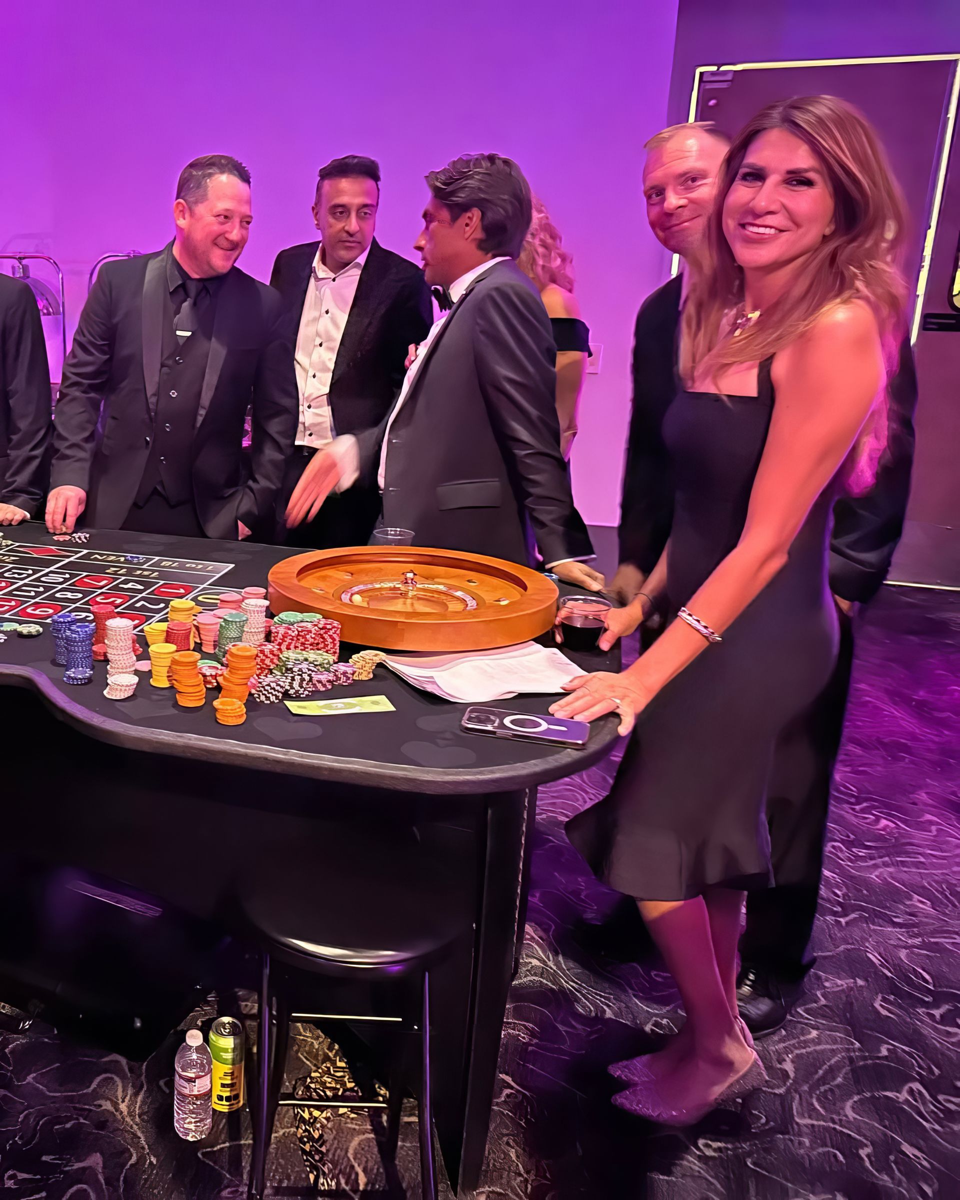 People gathered around a table with drinks and a large cake at a purple-lit event.