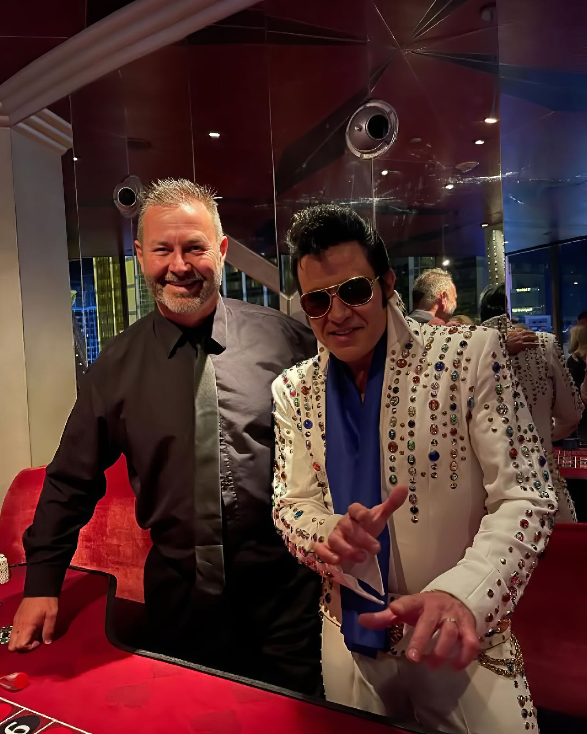 Two men pose at a red-lit venue, one in a black shirt and the other in a white studded jacket and sunglasses.