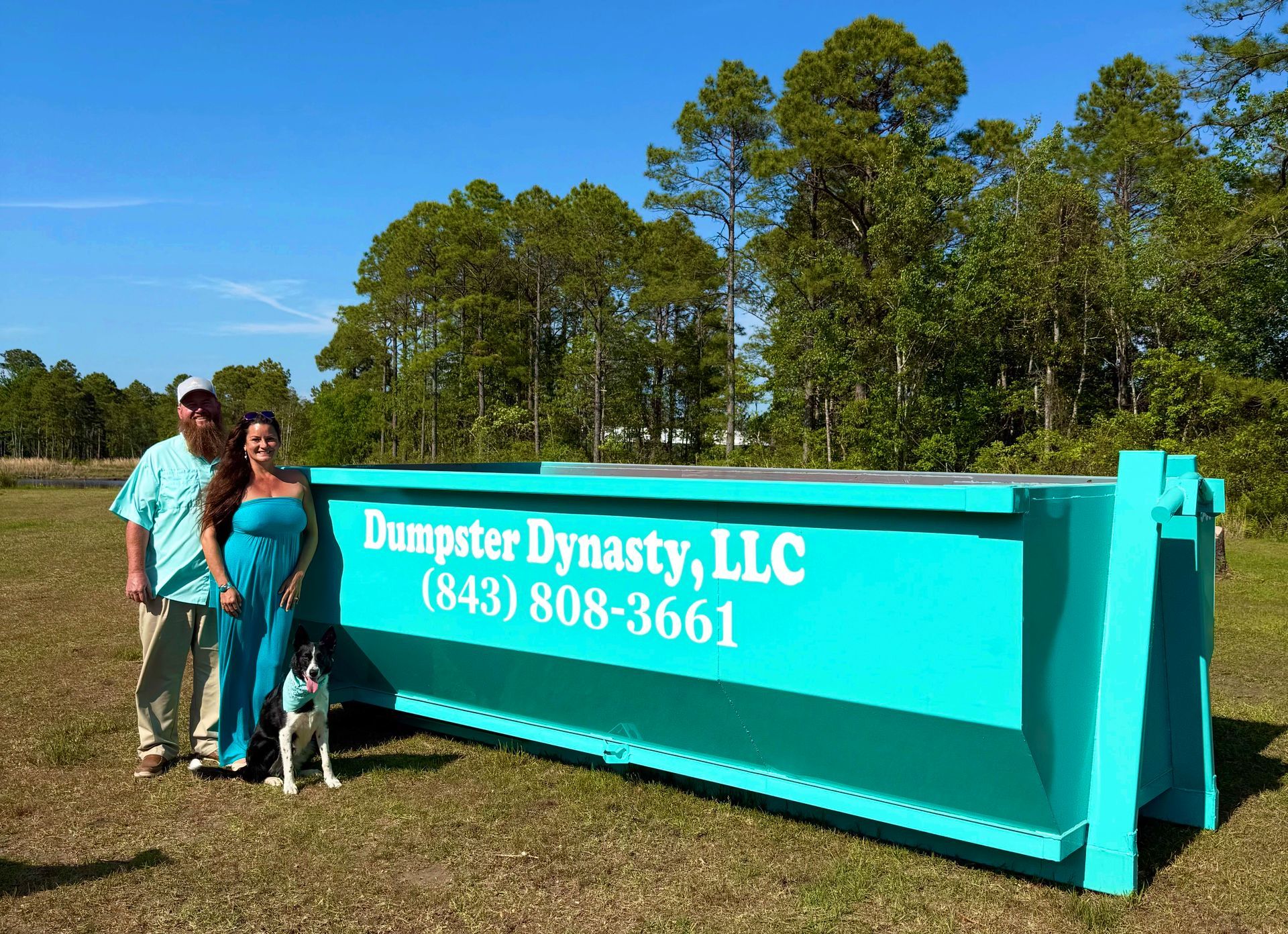 Two people standing beside a turquoise dumpster labeled “Dumpster Dynasty, LLC” in a grassy field.