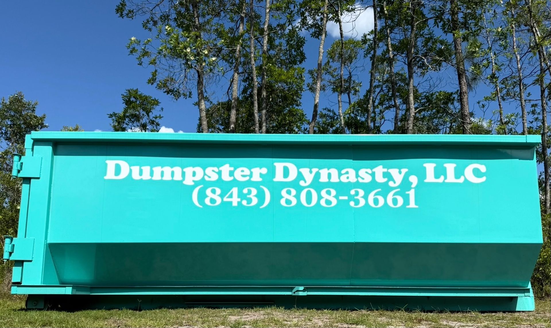 Turquoise Dumpster Dynasty LLC dumpster with phone number, outdoors against trees and blue sky