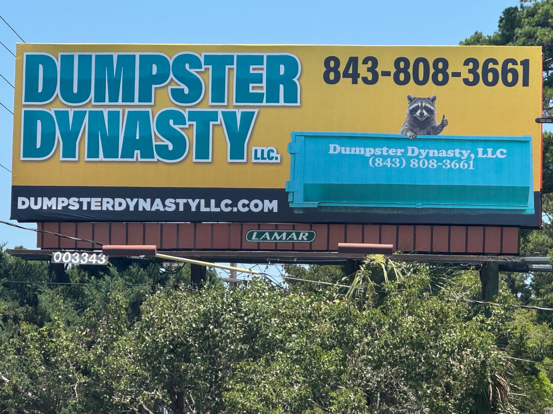 Dumpster Dynasty billboard with blue and yellow sign, phone number, and website above trees