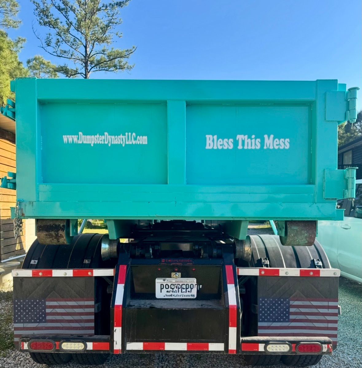 Turquoise dump truck with “Bless This Mess” on the back, viewed from behind on a road.