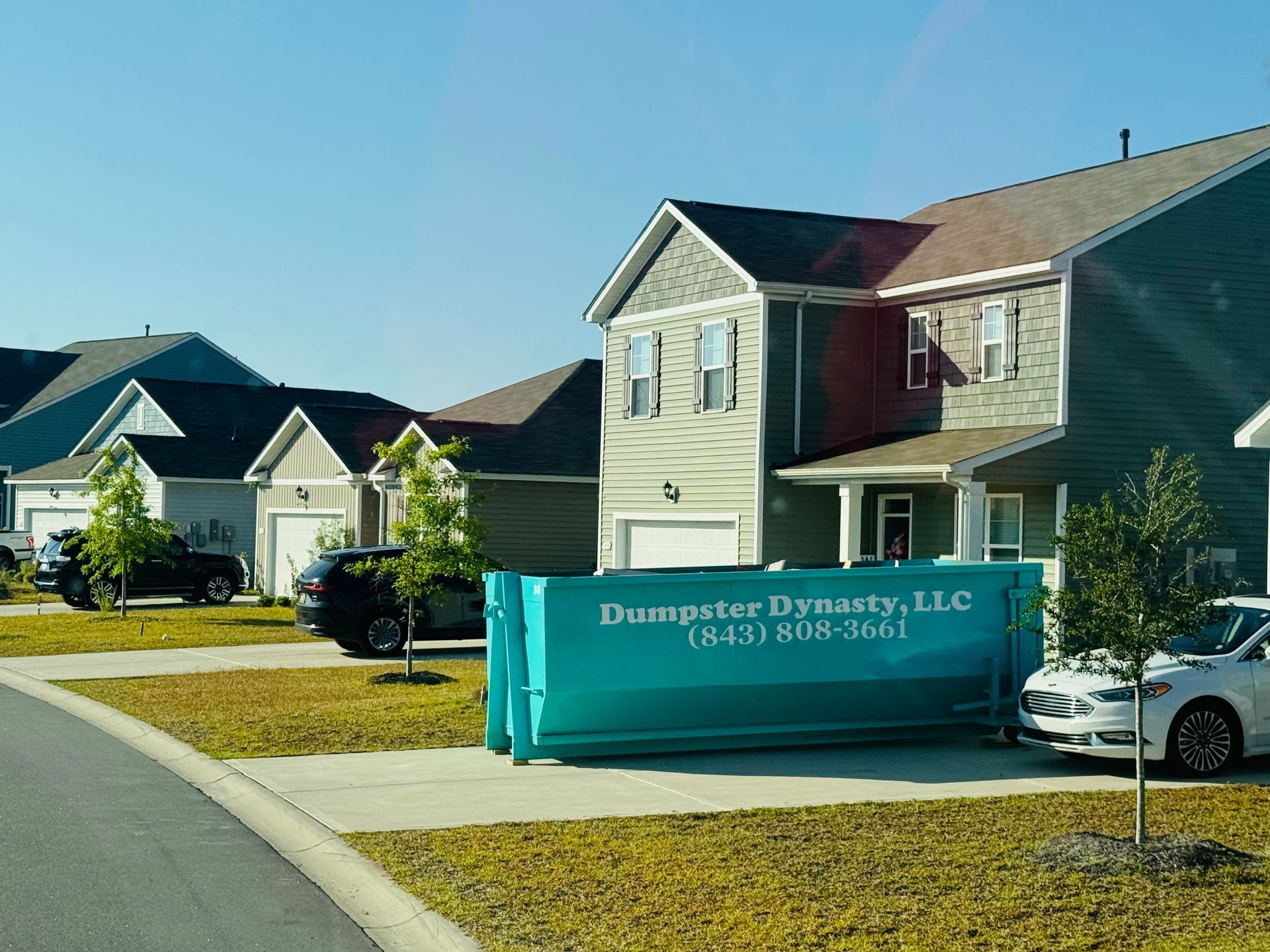 Suburban street with a turquoise dumpster bin in front of beige houses and a parked white car