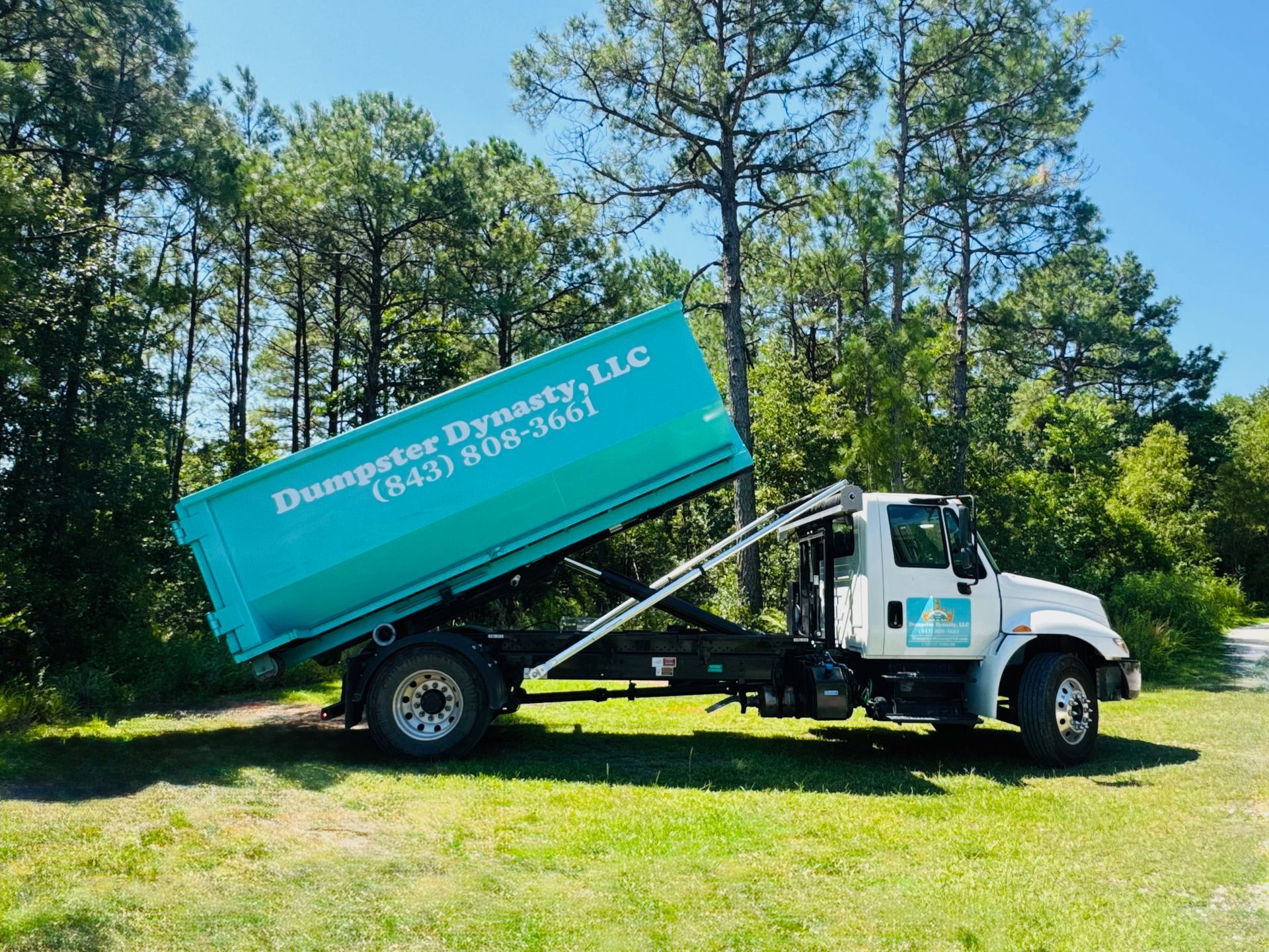 Turquoise dump truck tipping its bed on a grassy roadside near pine trees