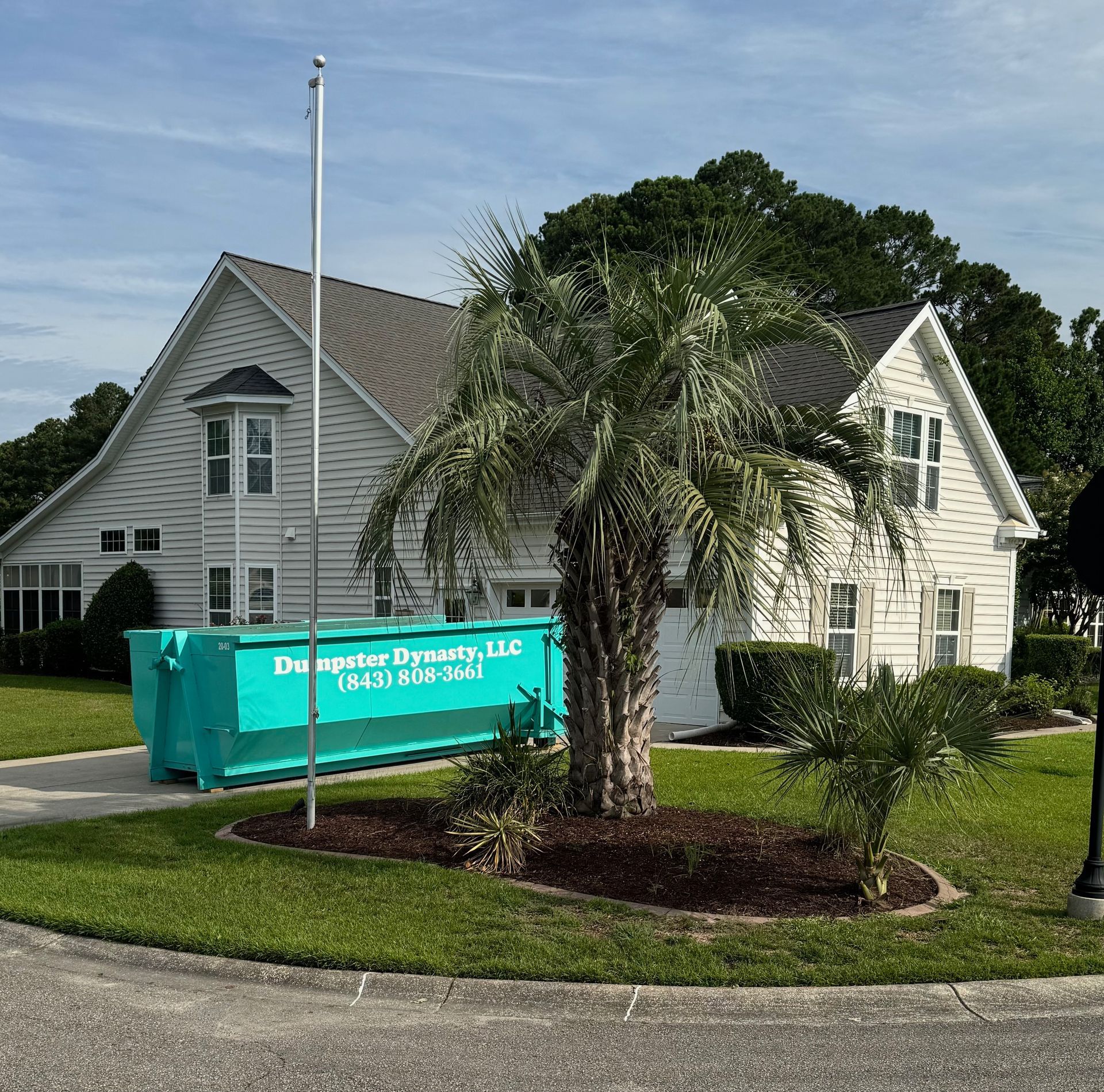 White house with palm tree, turquoise dumpster, and green lawn under a blue sky
