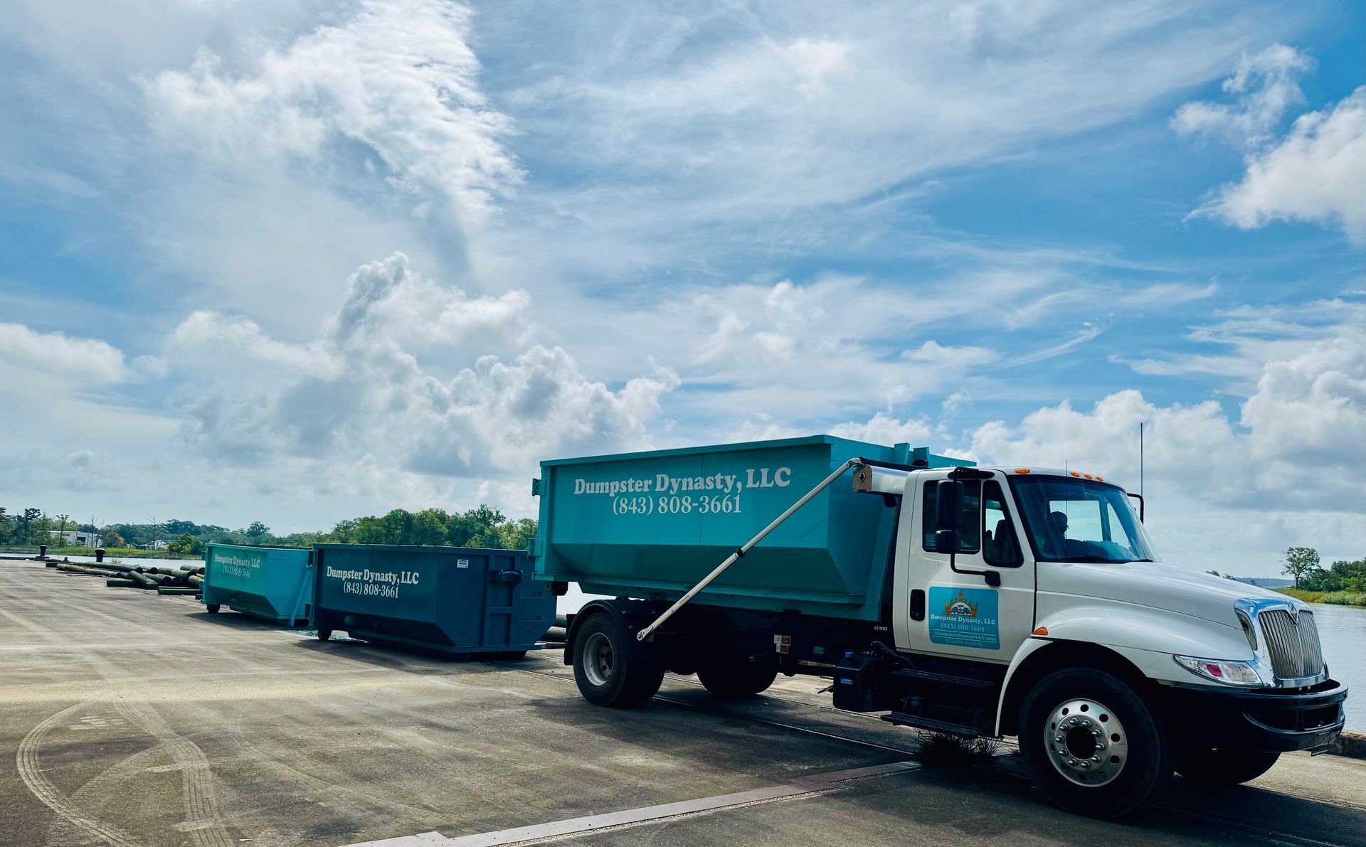 Blue trash truck towing green dumpsters on a road under a cloudy sky