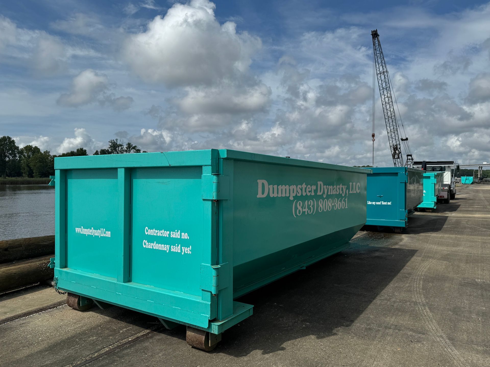 Turquoise dumpsters lined up on a paved waterfront dock under cloudy skies
