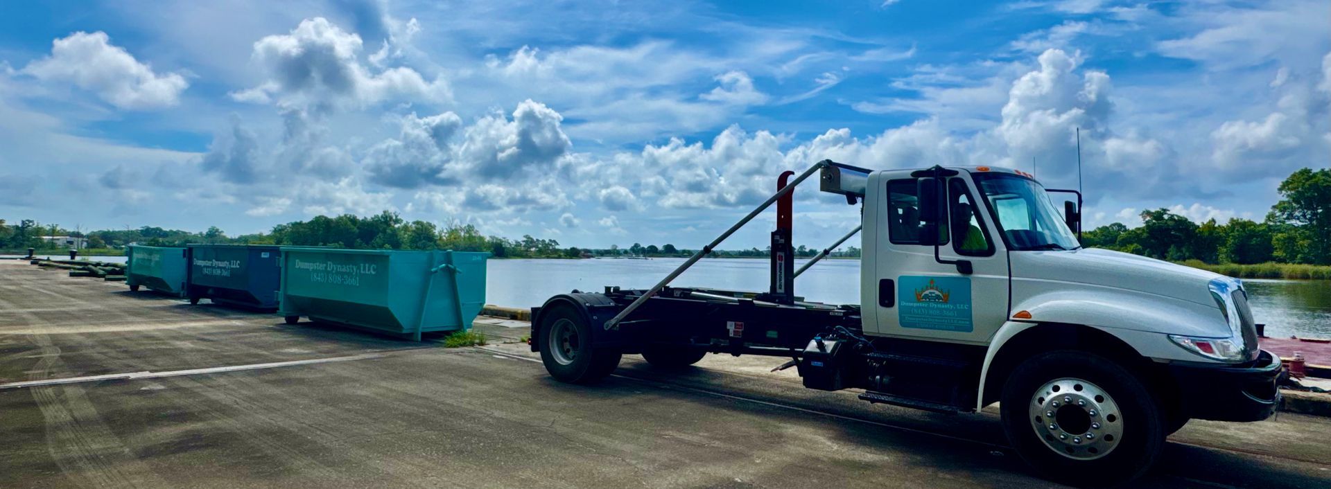 Tow truck hauling green dumpsters along a dirt lot under a cloudy sky