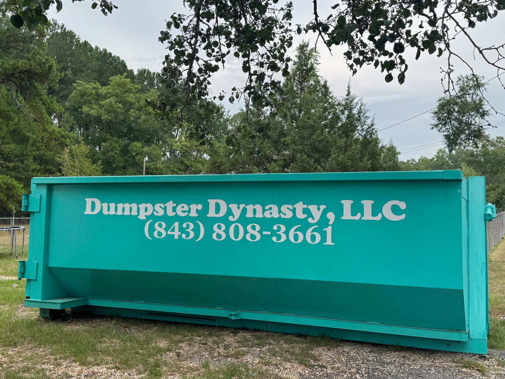 Teal Dumpster Dynasty LLC dumpster with phone number in a wooded outdoor setting