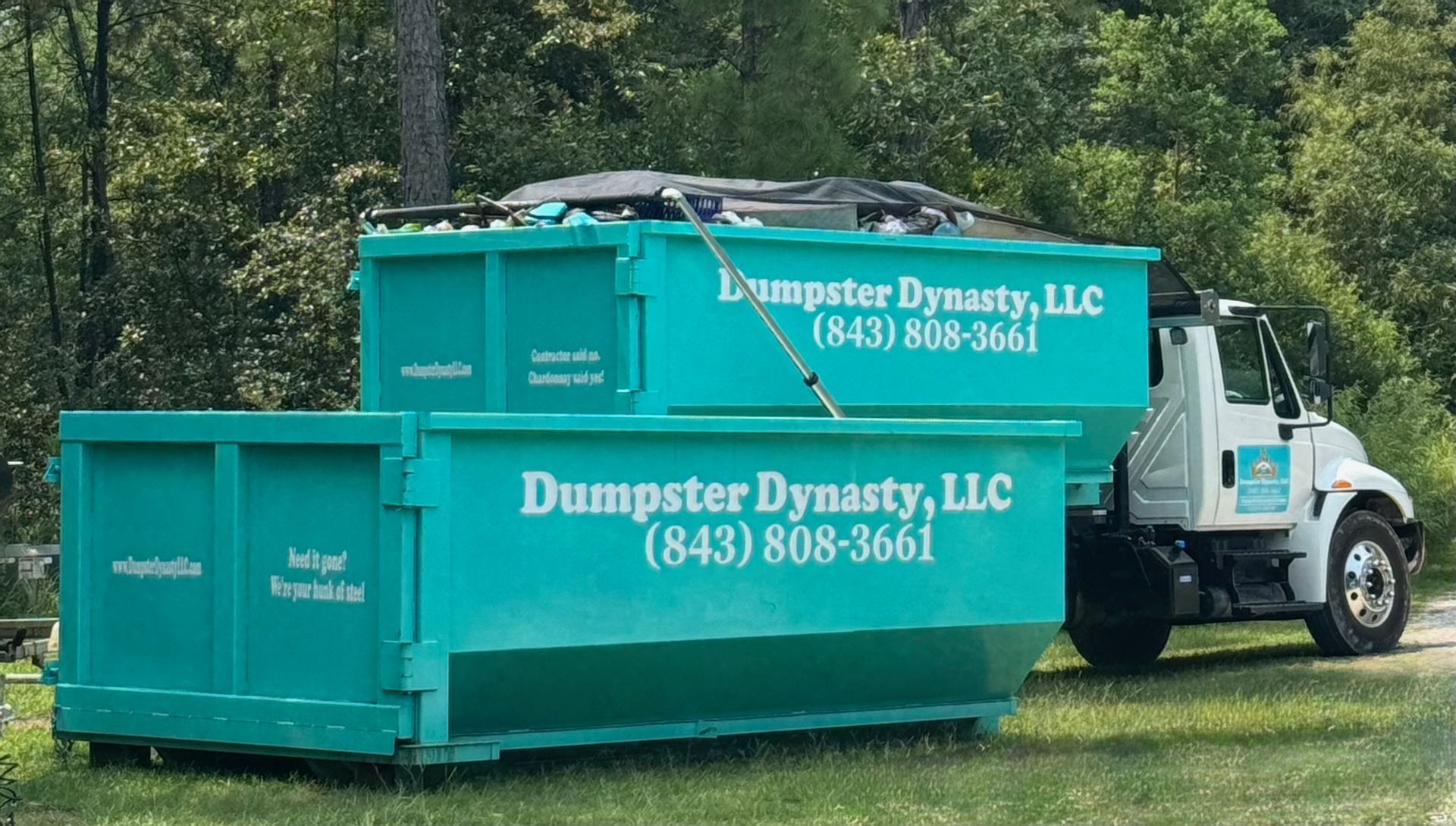 Green Dumpster Dynasty dumpster with logo and phone number beside a white truck in a grassy lot