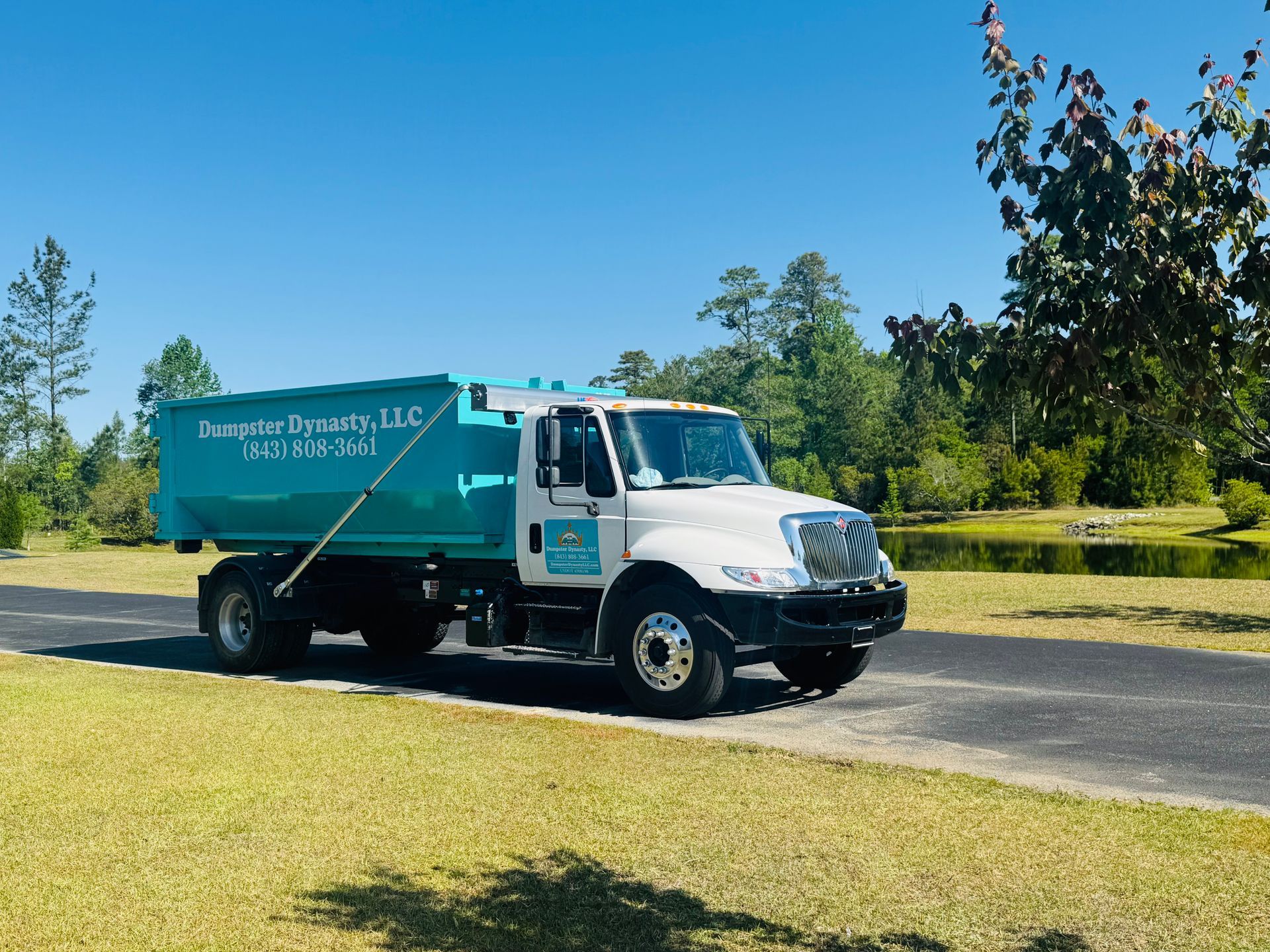 Turquoise waste truck parked on a driveway beside a grassy yard and trees under a blue sky