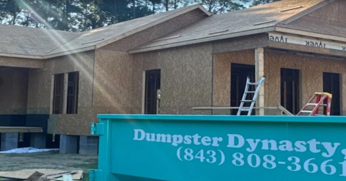 House under construction with a teal Dumpster Dynasty dumpster in front and ladders near the roof