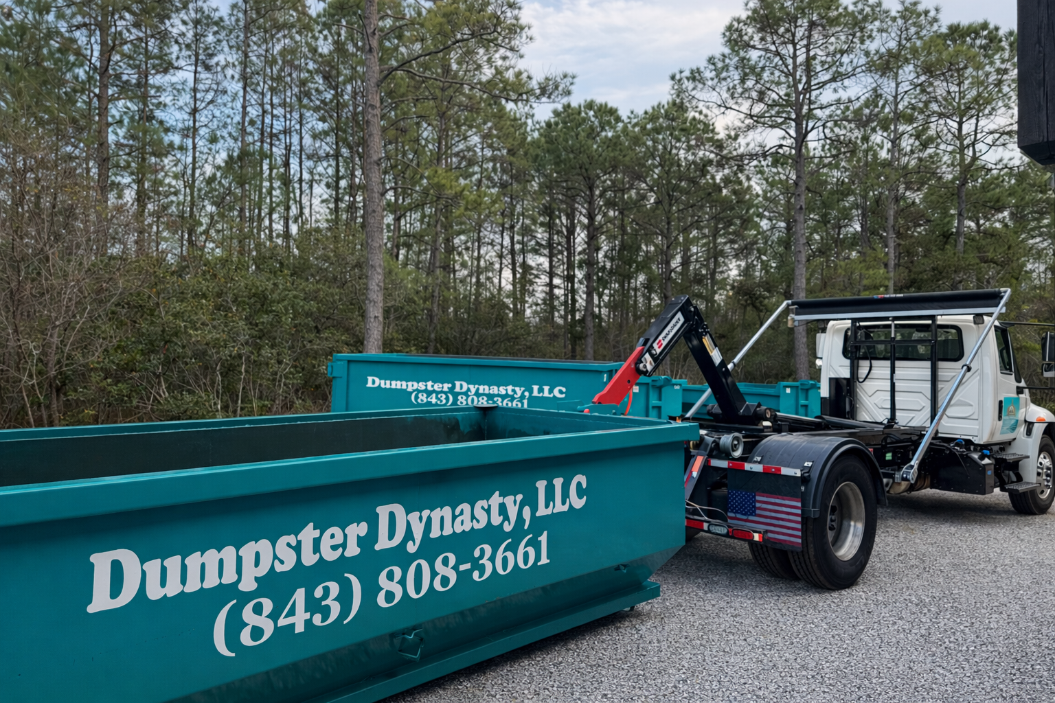 Teal Dumpster Dynasty LLC dumpster on trailer beside a white truck on a gravel road, wooded background