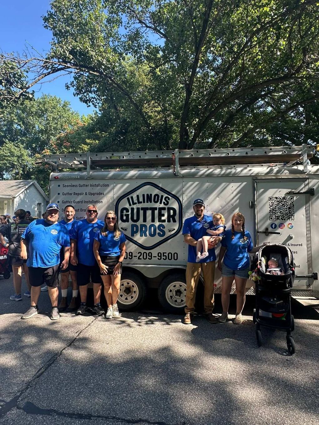 Illinois Gutter Pros team in blue shirts posing beside a service trailer on a sunny street