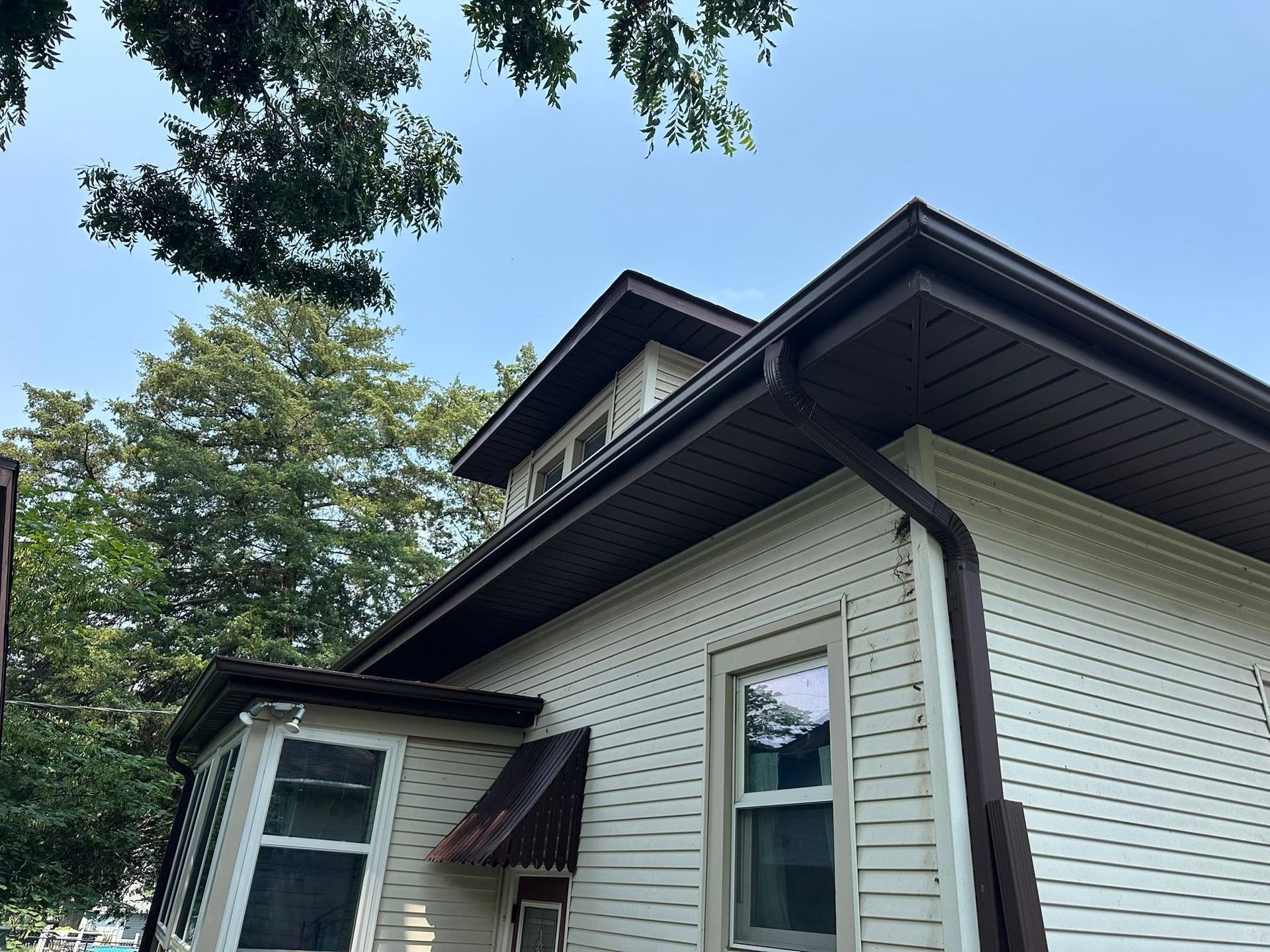 White house exterior with vinyl siding, dark roof, and trees overhead under a blue sky