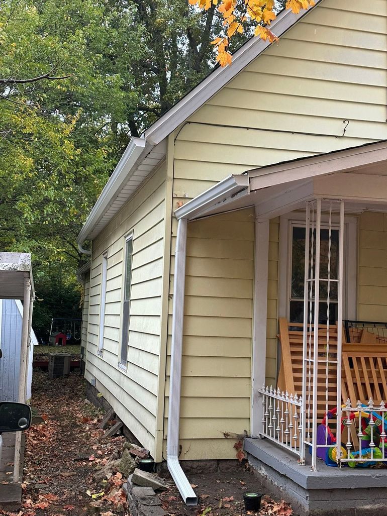 Side view of a beige house with white trim, downspout, and a small covered porch with white railing.