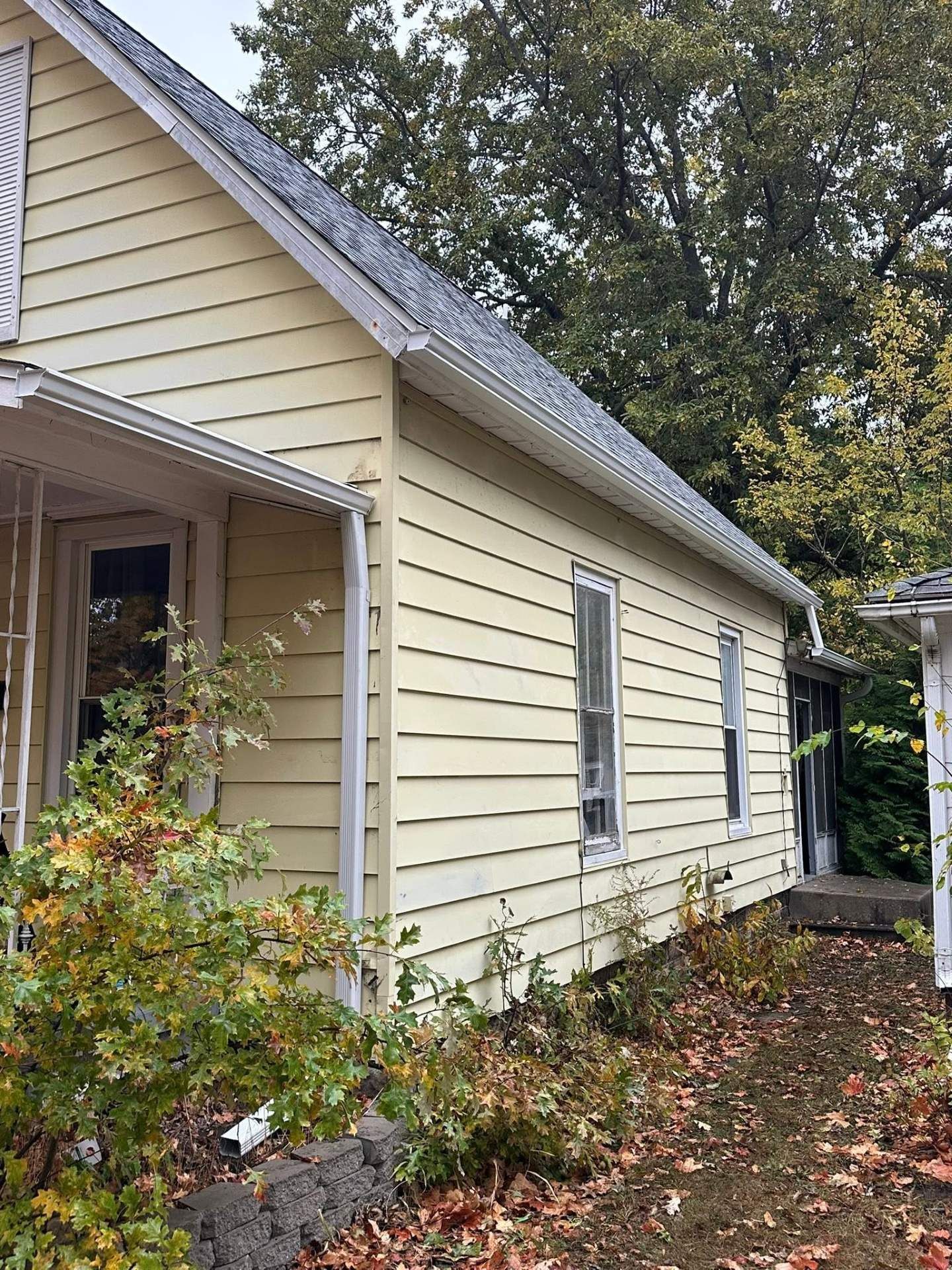 Yellow house exterior with white trim, side window, porch, and fallen autumn leaves along the foundation