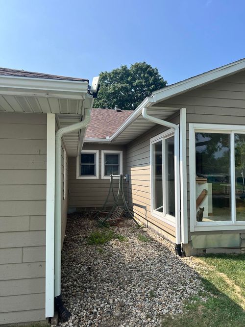 Narrow gravel path between two light-colored houses, with windows and a small roofed connector in a backyard.